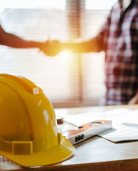 Accord pour un Projet de Construction et Casque Jaune Casque de chantier jaune au premier plan et poignée de main entre professionnels du bâtiment en arrière-plan flou.