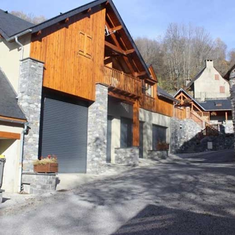 Maison alpine en bois et pierre avec garage moderne Chalet moderne en bois et pierre avec une porte de garage grise et un balcon dans un village de montagne.