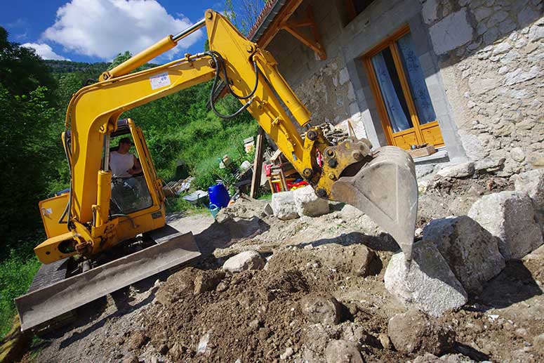 Mini pelle excavatrice : travaux de terrassement maison Mini pelle jaune déplaçant de grosses roches pour le terrassement devant une maison ancienne en pierre.