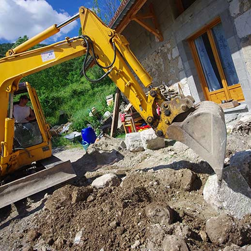 Mini-pelle en action pour travaux d'excavation de jardin Pelleteuse jaune en action pour des travaux d'excavation et d'aménagement près d'une maison en pierre.