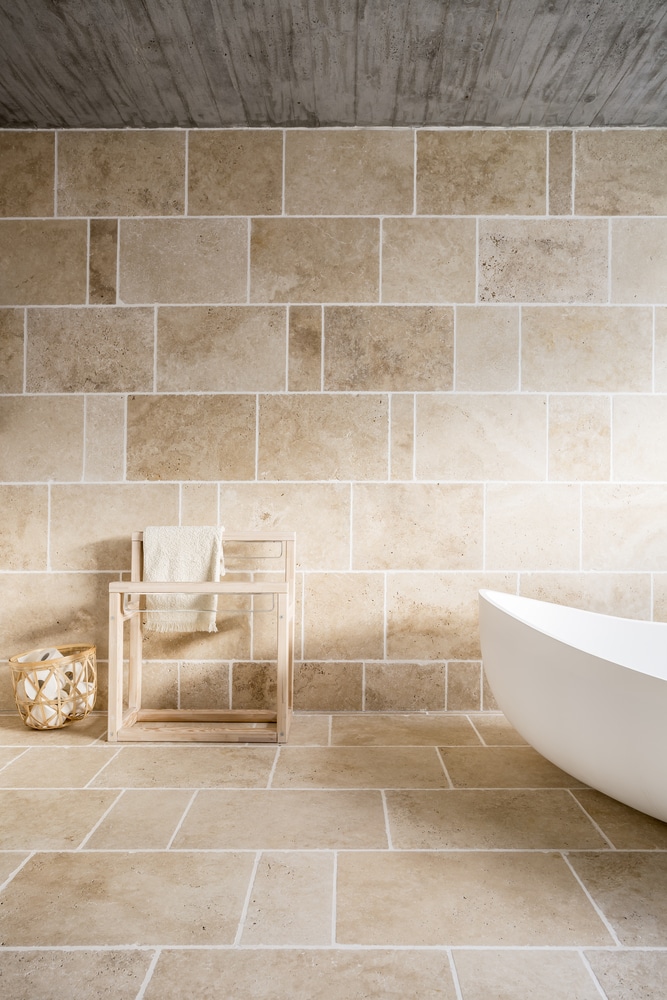 Salle de bain en pierre beige avec baignoire îlot design Salle de bain moderne avec carrelage en pierre beige, baignoire îlot blanche et porte-serviette en bois minimaliste.