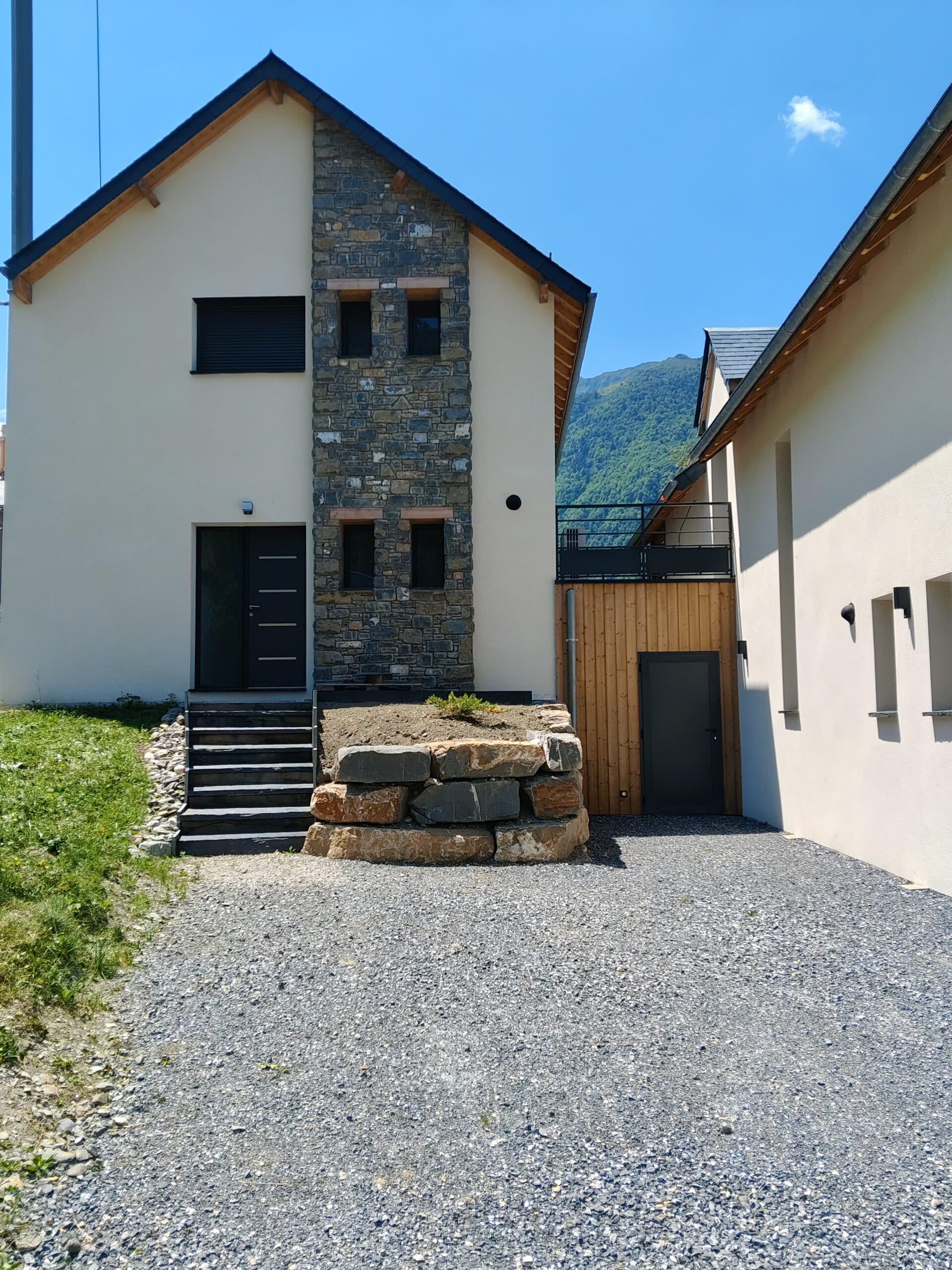 Maison moderne à façade en pierre et bois avec escaliers noirs et allée en gravier devant la montagne.